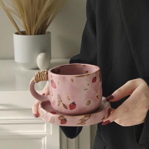 May include: Pink ceramic teacup and saucer set decorated with red strawberries. The teacup has a woven handle. The set is held by a person wearing a black shirt. A white vase with dried plants is in the background.