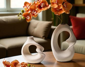 May include: Two off-white ceramic vases with a unique, open design sit on a wooden table. One vase holds orange orchids. The vases have a modern, sculptural aesthetic, adding a touch of elegance to the room.