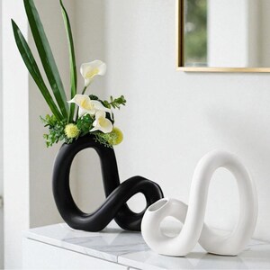 May include: A black, abstract ceramic vase with a unique looped design, holding green leaves, white calla lilies, and small green accents. A smaller white vase with a similar design sits beside it. The vases are on a white marble surface.