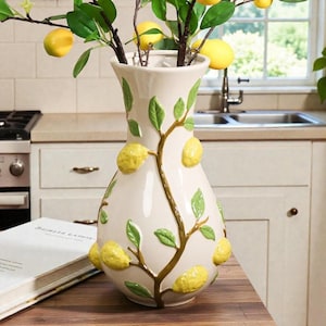 May include: A cream ceramic vase featuring a lemon and leaf design. The vase has a wide base and a narrow neck, with yellow lemons and green leaves decorating the surface. The vase is filled with lemon branches.