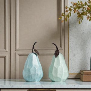 Puede incluir: Dos esculturas decorativas de pera. Una es azul claro, la otra verde claro. Ambas tienen un diseño facetado y un tallo marrón oscuro con una hoja. Se exhiben sobre una encimera blanca.