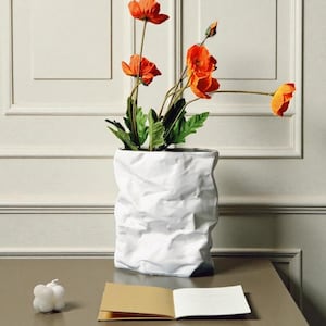 May include: A white, crumpled paper bag-style vase holding orange poppies with green leaves. A small, white bubble candle and a small open notebook sit on a brown surface. The vase is the focal point.