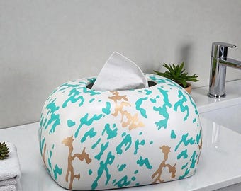 May include: A white tissue box with a rounded shape, decorated with a teal and gold splatter pattern. A white tissue is pulled from the top opening. The box is on a white countertop near a sink.