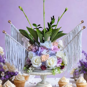 May include: A floral arrangement with white, purple, and lavender flowers on a white cake stand. The cake is decorated with white roses and greenery. White fringe banners are attached to the cake. Cupcakes with white frosting are in the foreground. The text "GIFTS BY KNJ" is in the upper right corner.
