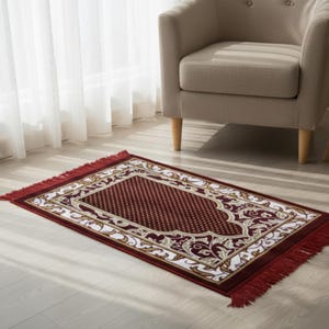 May include: A rectangular prayer rug with a burgundy field and a white and gold border featuring floral designs. The rug has a fringed edge and is placed on a light-colored wooden floor.