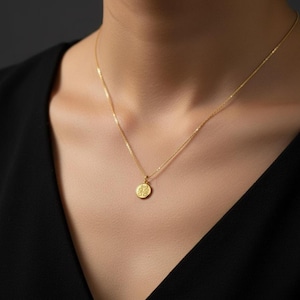 May include: A gold necklace with a small, circular pendant displaying a detailed pattern. The pendant is suspended from a slender chain, offering a refined appearance. The necklace's gold colour is accentuated by the dark backdrop.