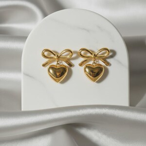 May include: A pair of gold earrings, each with a bow design and a heart-shaped pendant. The earrings are displayed on a white marble surface, with a white satin fabric in the background. The earrings are made of a gold-colored metal.
