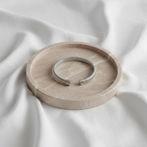 Peut inclure: Un bracelet manchette ouvert argenté avec un design côtelé repose sur un plateau rond en marbre beige. Le bracelet a une esthétique moderne et minimaliste. Le plateau est posé sur un tissu blanc texturé.