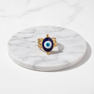 May include: A gold ring with a dark blue oval centrepiece, featuring a white and light blue eye design. The ring has three bands and small gold accents. It rests on a white marble surface.