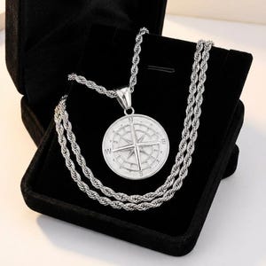Puede incluir: Un collar con colgante de brújula plateada con una cadena de cuerda, presentado en una caja de joyería de terciopelo negro. El colgante presenta un diseño detallado de rosa de los vientos con marcadores direccionales. Un accesorio elegante.