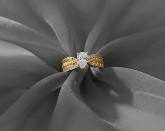 May include: A gold ring featuring a clear, pear-cut gemstone. The ring's band has an intricate, textured pattern. The ring is displayed on a grey fabric background.