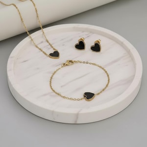 May include: A gold-coloured jewellery set comprising a necklace, bracelet, and earrings, each with a black heart-shaped pendant. The set is presented on a white marble tray against a grey background. The earrings are studs.