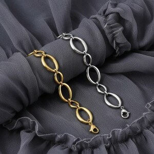 Könnte beinhalten: Zwei Armbänder mit ovalen Kettengliedern, eines goldfarben und eines silberfarben, werden auf einem grauen Stoff präsentiert. Jedes Armband hat einen Karabinerverschluss. Das goldfarbene Armband befindet sich links, das silberfarbene rechts.