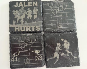 May include: Four square black slate coasters with etched football-themed designs. One coaster features the name "JALEN HURTS" with football players. Other coasters show football plays and the text "Philly Special PATRIOTS".