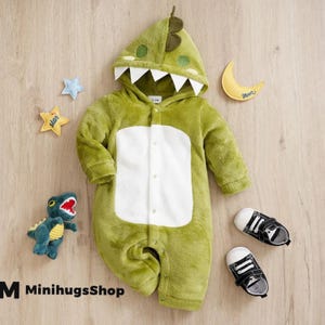 Puede incluir: Un mono verde de bebé con forma de dinosaurio, con capucha con dientes blancos y una pequeña aleta. El mono tiene una barriga blanca y se abrocha con botones. Un par de zapatos de bebé blancos y negros están al lado.