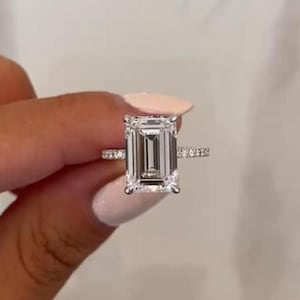 May include: A silver ring with a large, rectangular, emerald-cut diamond as the centerpiece. The band is adorned with smaller, sparkling stones. The ring is held up against a neutral background, showcasing its brilliance and design.