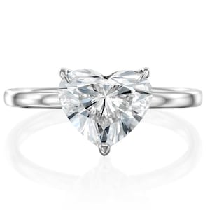 May include: A silver ring featuring a large, heart-shaped diamond. The diamond is clear and faceted, catching the light. The band is smooth and polished, with a simple design. The ring is set against a white background.