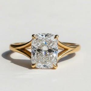 Op de afbeelding: Een gouden ring met een grote, rechthoekige, heldere edelsteen. De band heeft een split-schacht ontwerp aan weerszijden van de edelsteen. De ring staat tegen een witte achtergrond.