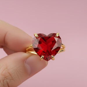 May include: A gold ring featuring a large, heart-shaped, faceted, deep red gemstone. The ring has a double band design and is held against a soft pink background. The gemstone is the focal point, catching the light and showcasing its brilliant cut.