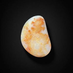 Puede incluir: Una gema pulida de forma ovalada, con una base blanca crema y patrones intrincados de naranja y amarillo. La piedra tiene una forma suave y redondeada, y los colores se mezclan en un diseño natural. La gema está sobre un fondo negro.