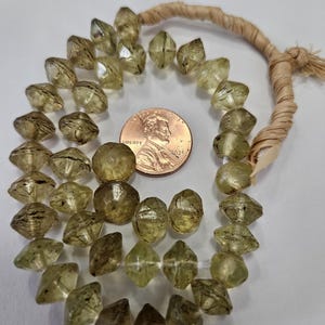 May include: A strand of faceted, translucent, pale olive-green beads with dark internal inclusions, arranged in a circular pattern next to a copper-coloured United States penny for scale. The beads are strung on a light-coloured, fibrous cord.