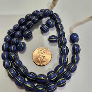 May include: A strand of oval-shaped dark blue glass beads featuring vertical yellow stripes, arranged in a circular pattern next to a copper-colored United States one-cent coin for scale, threaded on a light-colored string.