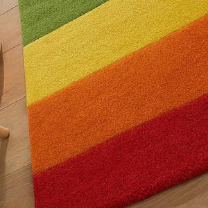 K&ouml;nnte beinhalten: Nahaufnahme eines farbenfrohen Teppichs mit diagonalen Streifen in Gr&uuml;n, Gelb, Orange und Rot. Der Teppich hat eine strukturierte Oberfl&auml;che und liegt auf einem Holzboden. Das Design des Teppichs verleiht dem Raum eine lebendige Note.
