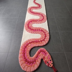 Könnte beinhalten: Ein langer, rechteckiger Läufer mit cremefarbenem Hintergrund und einem großen, geschwungenen Schlangenmotiv. Die Schlange besteht aus verschiedenen Rosa-, Korallen- und Rottönen und hat eine texturierte, getuftete Oberfläche sowie eine rote gespaltene Zunge.