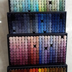 K&ouml;nnte beinhalten: Eine Pr&auml;sentation von Teppichfarbmustern in rechteckigen Tabletts. Die Muster bestehen aus kleinen, flauschigen Pompons, die eine Reihe von Farben zeigen, von tiefen Rot- und Blaut&ouml;nen bis hin zu sanften Pastellt&ouml;nen und leuchtenden Oranget&ouml;nen.