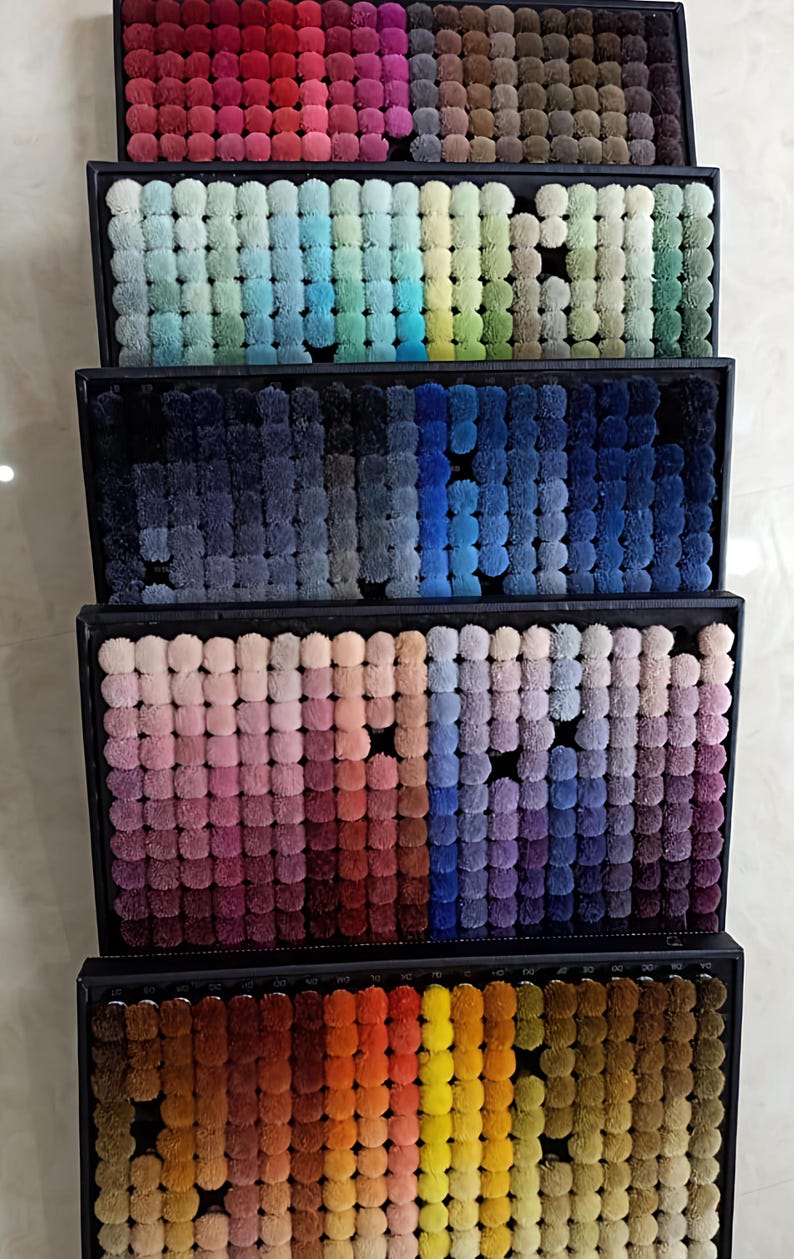 K&ouml;nnte beinhalten: Eine Pr&auml;sentation von Teppichfarbmustern in rechteckigen Tabletts. Die Muster bestehen aus kleinen, flauschigen Pompons, die eine Reihe von Farben zeigen, von tiefen Rot- und Blaut&ouml;nen bis hin zu sanften Pastellt&ouml;nen und leuchtenden Oranget&ouml;nen.