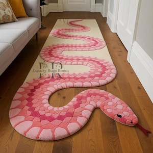 Peut inclure: Un tapis en forme de serpent rose et rouge sur fond beige. Le tapis est fait d'une matière douce et texturée et présente un motif détaillé. La tête du serpent a une langue rouge et des yeux noirs. Le tapis est posé sur un sol en bois.
