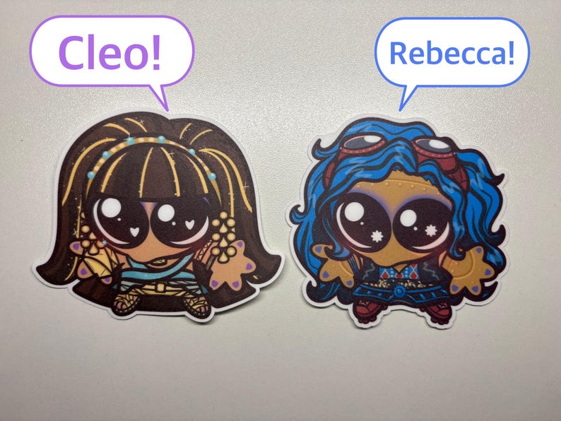 Op de afbeelding: Twee gestanste stickers met gestileerde stripfiguren met grote ogen. De linker sticker toont een personage met donker haar en de tekst Cleo! in een tekstballon. De rechter sticker toont een personage met blauw haar en de tekst Rebecca! in een tekstballon.