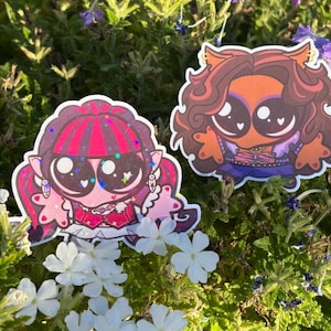 Op de afbeelding: Twee gestanste stickers met gestileerde stripfiguren met grote ogen. De linker sticker toont een personage met een roze huid, donker haar met roze highlights en een rode outfit. De rechter sticker toont een personage met bruin haar en een oranje outfit.