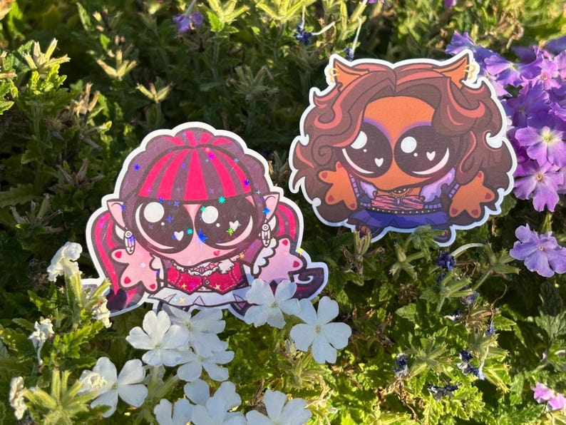 Op de afbeelding: Twee gestanste stickers met gestileerde stripfiguren met grote ogen. De linker sticker toont een personage met een roze huid, donker haar met roze highlights en een rode outfit. De rechter sticker toont een personage met bruin haar en een oranje outfit.