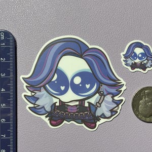 Op de afbeelding: Een gestanste sticker met een gestileerd personage met grote blauwe ogen, paars en blauw haar en een donkere outfit, naast een liniaal van ongeveer 7,5 cm hoog en een munt voor schaal, inclusief een kleinere bijpassende sticker.
