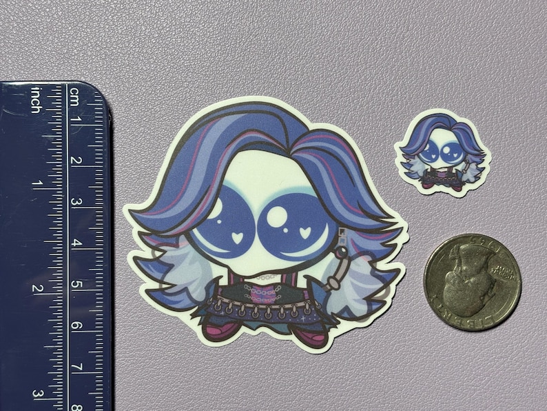 Op de afbeelding: Een gestanste sticker met een gestileerd personage met grote blauwe ogen, paars en blauw haar en een donkere outfit, naast een liniaal van ongeveer 7,5 cm hoog en een munt voor schaal, inclusief een kleinere bijpassende sticker.