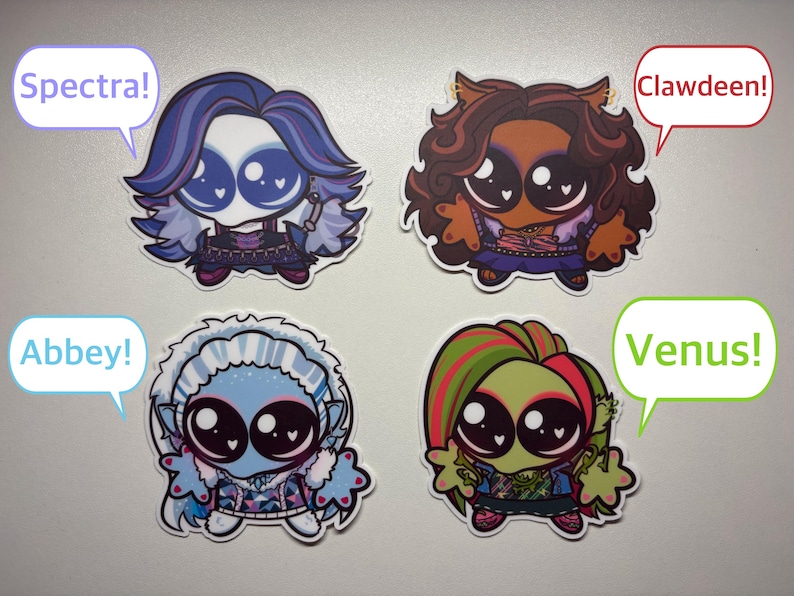 Op de afbeelding: Vier chibi-stijl karakterstickers met grote ogen en hartvormige reflecties. Links boven een figuur met paars haar en een tekstballon "Spectra!". Rechts boven bruin haar en "Clawdeen!". Links onder blauw haar en "Abbey!". Rechts onder groen haar en "Venus!".