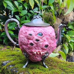 May include: A pink, grotesque teapot with multiple eye-like features and a toothy grin. The teapot has a silver spout, handle, and lid, and sits on three small silver feet. The background features lush green foliage and moss.