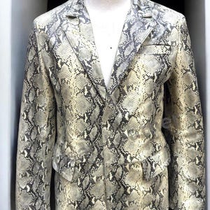 May include: A blazer featuring a prominent snakeskin pattern in shades of cream, beige, and dark gray. The garment includes a notched lapel, long sleeves, and two front flap pockets. The design is consistent throughout the front of the jacket.