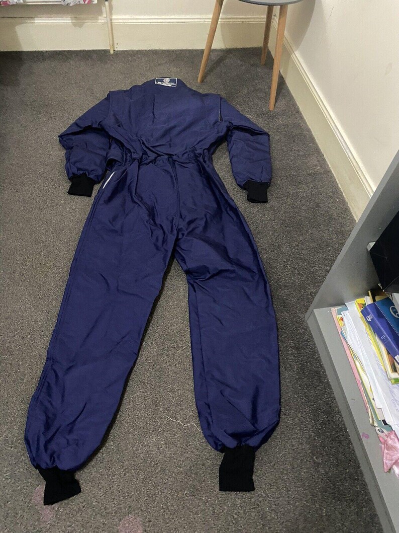 May include: A navy blue racing suit with black cuffs and collar. The one-piece garment has long sleeves and legs, with a white stripe detail. The suit is lying on a gray carpet.