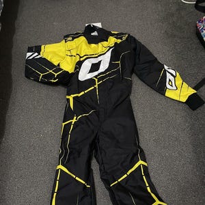 Pu&ograve; includere: Tuta da corsa nera e gialla con dettagli bianchi. La tuta presenta un design geometrico con linee gialle e un grande logo bianco sul petto. Le maniche e le spalle sono principalmente gialle, con polsini neri.