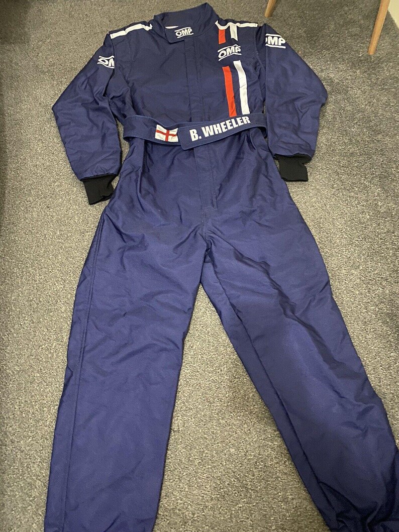 May include: A navy blue racing suit with white and red stripes on the chest. The suit has the name "B. Wheeler" on the belt and the OMP logo. The suit has black cuffs and white stripes on the shoulders.