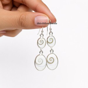 Op de afbeelding: Een paar zilveren oorbellen met twee ovale Shiva-oog schelp cabochons per oorbel. De witte schelpen vertonen een natuurlijk spiraalpatroon in bruine en grijze tinten, gezet in gepolijste zilveren monturen met haaksluitingen voor aan de oren.