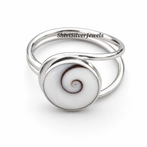 Puede incluir: Un anillo de plata con una incrustación circular de concha de ojo de Shiva con un patrón de espiral marrón natural. La banda está formada por dos finos hilos de plata pulida que se unen en el engaste. El texto ShiviSilverJewels es visible.