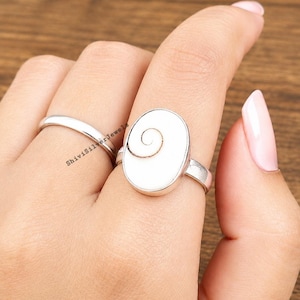 Puede incluir: Un anillo de plata con una piedra ovalada blanca que presenta un patrón de espiral marrón natural, acompañado de un anillo de banda de plata liso. El anillo ovalado tiene un engaste de metal liso y un acabado pulido con un diseño de espiral.