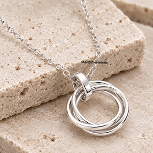 May include: A silver-toned necklace featuring a pendant composed of three interlocking circular rings suspended from a delicate chain. The metal has a polished, reflective finish. The text ShiviSilverJewels is visible near the pendant attachment.