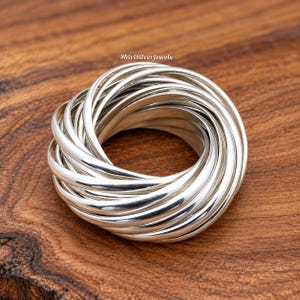 May include: A multi-band ring composed of numerous thin, polished silver-coloured circular bands intertwined to form a single cohesive unit. The text ShiviSilverJewels is visible in small black font above the ring, which features a smooth, reflective surface.