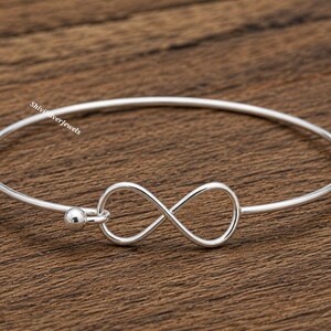 Może przedstawiać: Srebrzysta bransoletka typu bangle z centralnym symbolem nieskończoności i małym kulistym koralikiem na jednym końcu drutu. Napis ShiviSilverJewels jest wydrukowany na biało po lewej stronie cienkiej, polerowanej metalowej obręczy.