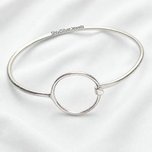 May include: A silver bangle bracelet featuring a circular design at the centre. The bracelet has a simple, elegant design and is made of a shiny metal. The text "ShiviSilverJewels" is printed on the bracelet.
