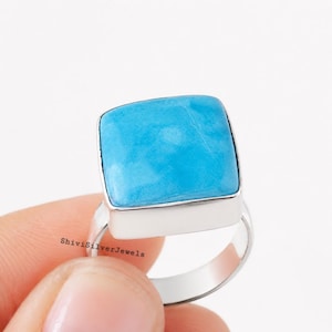 May include: A silver ring featuring a large, square-shaped, smooth, bright turquoise-colored stone set in a polished metal bezel. The band is plain and metallic. The text ShiviSilverJewels is printed in small black letters on the side of the ring.